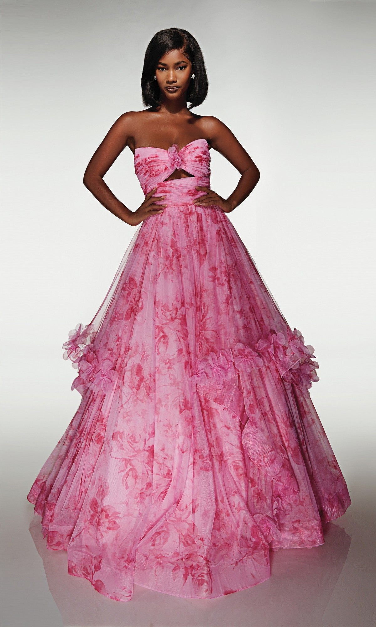 A woman stands in the Alyce 62042 long prom dress, a strapless pink floral ballgown featuring a fitted bodice with cut-out detail and a full ruffle skirt adorned with layered ruffles.